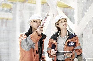 The picture shows 2 people in work clothes walking around a construction site and taking notes